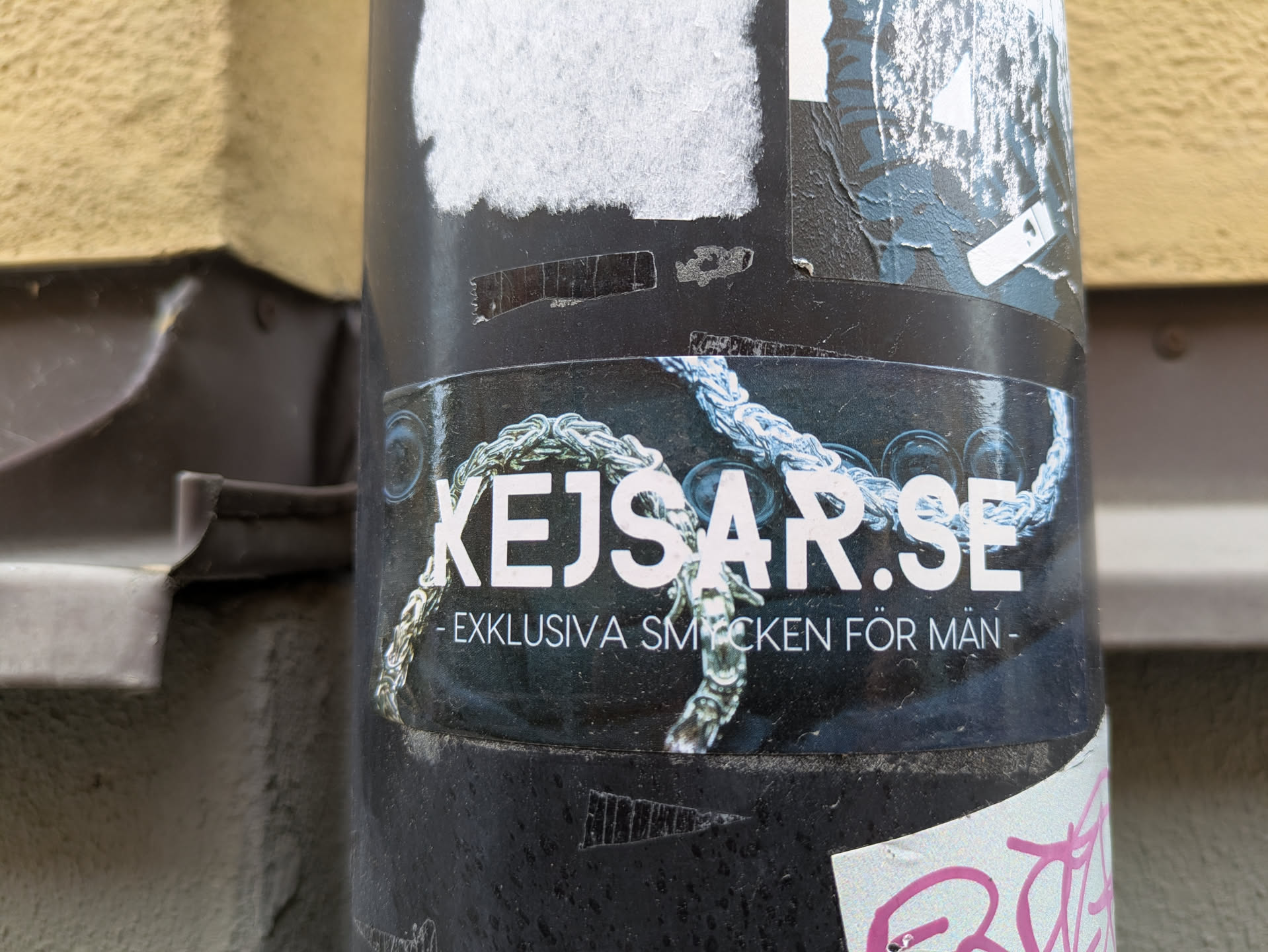 The sticker is predominantly dark with white lettering. Prominently displayed in large white font is 'KEJSAR.SE'. Below this, in smaller white text, reads '- EXKLUSIVA SMYCKEN FÖR MÄN -'. The lettering is overlaid on a dark background that features a detailed image of a thick, intertwined silver-colored chain with blueish highlights, suggesting luxury and craftsmanship. The sticker is slightly curved, conforming to the cylindrical surface it's adhered to, and shows some signs of wear, with minor peeling and scuffing at the edges and surface.