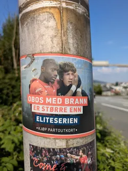 The sticker prominently displays two male football players, one with dark skin wearing a red jersey (likely SK Brann's colors) and another with light skin and curly hair appearing emotional or distressed, with his arm around the first player. The background is blurred, suggesting a stadium or field. Overlaying the image are large white and red text boxes. The text translates to 'OBOS with Brann is bigger than Eliteserien' and 'Buy season ticket'. The sticker is affixed to a weathered metal pole next to a road, with greenery on the left and a distant urban landscape on the right.