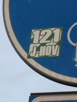 The sticker is rectangular with slightly rounded corners, featuring a solid dark green background. Prominently displayed in white, bold, blocky, sans-serif font with a dark outline are the characters '121' on the top line and 'J-HOV' directly below it. The overall style is clean and impactful, resembling a sports jersey number or a team identifier, contrasting sharply with the blue sign it's adhered to.