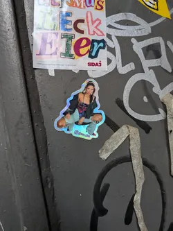 Street sticker A die-cut sticker featuring a photographic cutout of a young woman crouching forward with wide knees. She is wearing a dark off-the-shoulder jacket, a multicolored top, pale blue flared pants with cutouts, and red heels, with headphones around her head. The sticker has a shiny, holographic outline, and at the bottom edge, it displays an Instagram camera icon alongside the username '@zoe.zita'.