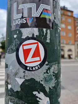 A round black and white sticker with a red Z in the center and the word "K L A R T" at the bottom. The sticker is on a green pole with other stickers around it.