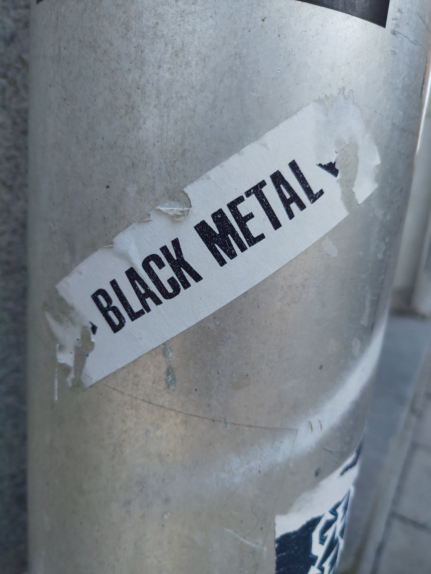 A simple sticker with the words "Black Metal" written in a bold, sans-serif font. The sticker is white with black text and is affixed to a grey metal pole. The sticker is slightly damaged around the edges.