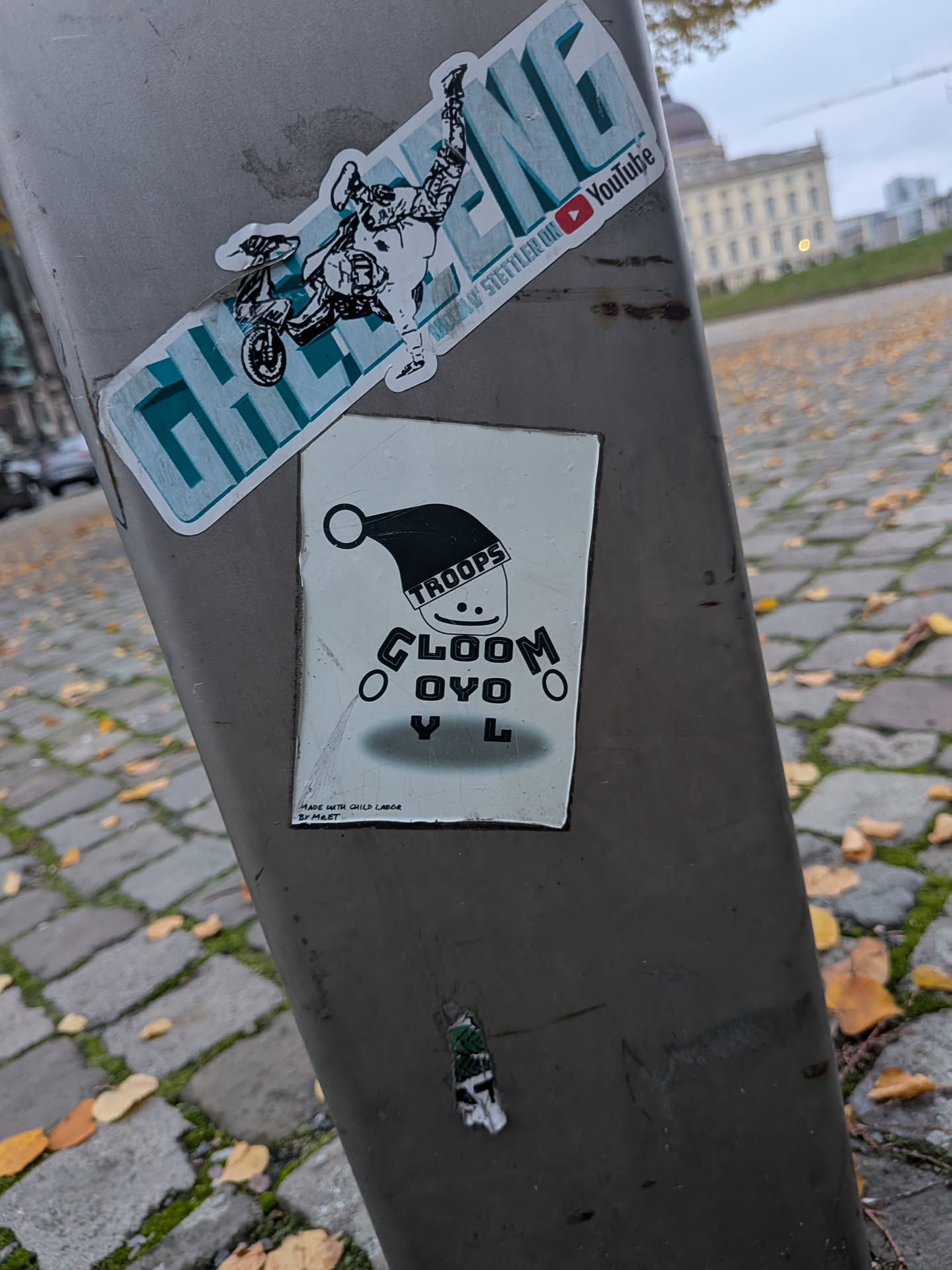 A rectangular white sticker featuring a black-and-white stylized character design. The figure has a simple smiley face wearing a black folded Santa-style cap with 'TROOPS' printed along the brim. Below the head, the torso consists of the word 'GLOOM' arched over 'O Y O' with small loop hands and the letters 'Y L' acting as legs, casting an oval shadow onto the ground. In the bottom-left corner, small printed text reads 'MADE WITH CHILD LABOR BY MRET'.