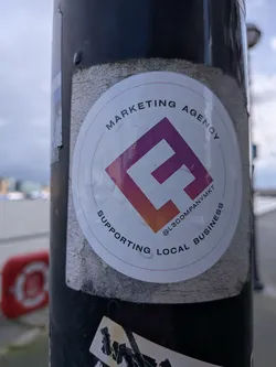 A circular white sticker adhered to a dark pole, featuring a central logo with a stylized, blocky 'L' or 'F' shape rendered in a purple-to-orange gradient. A thin, light brown or orange-red circle outlines the inner content. Arched text above the logo reads "MARKETING AGENCY", and below it, "SUPPORTING LOCAL BUSINESS". A social media handle, "@L3COMPANYMKT", is positioned horizontally below the logo. The sticker shows signs of outdoor exposure, with several small water droplets visible on its surface and some wear around its edges.