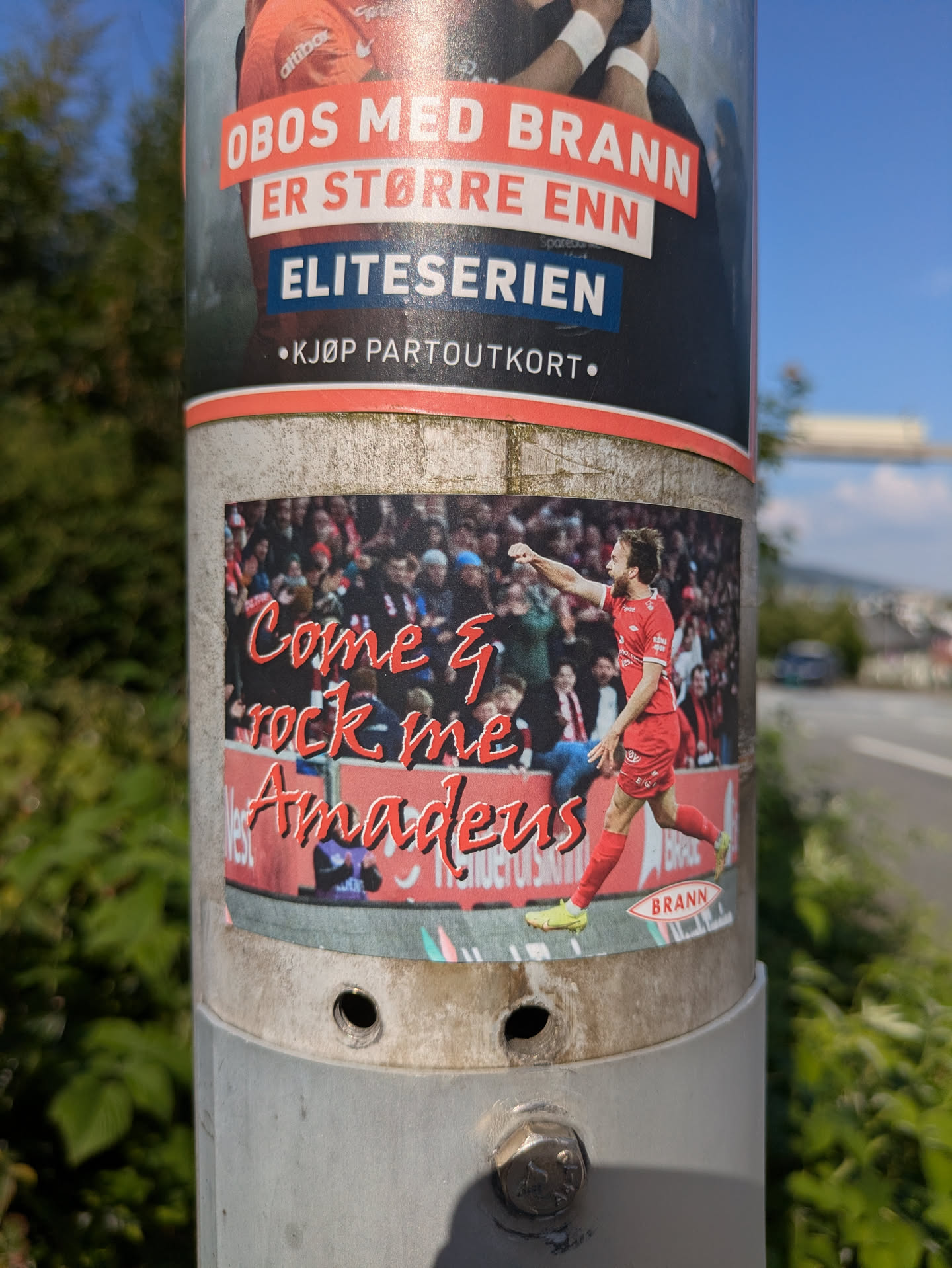 The sticker prominently displays a football (soccer) player from SK Brann, dressed in a red uniform, in what appears to be a celebratory or action pose during a match, with a blurry stadium crowd in the background. The text 'Come & rock me Amadeus' is overlaid in a vibrant red script font, referencing the famous song by Falco. At the bottom right of the sticker, there's a small red oval with the word 'BRANN', clearly identifying the team. The sticker is affixed to a utility pole, with another larger poster above it also related to Brann, mentioning 'OBOS' and 'Eliteserien'.