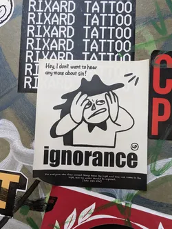 A black and white sticker with a cartoon character covering its ears and the word "ignorance" in bold text.  A quote about sin is included at the bottom. 