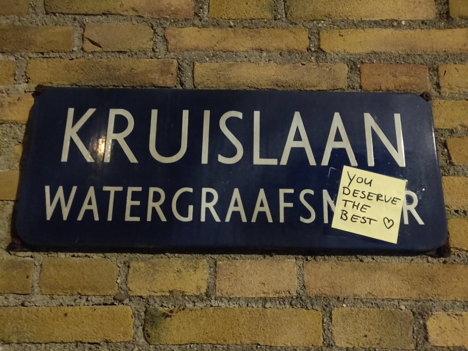 A yellow sticky note with the handwritten message "You deserve the best" stuck to a vintage blue street sign. The street sign reads "Kruislaan Watergraafsmeer" in white lettering. The photo was likely taken at night or in low light conditions.