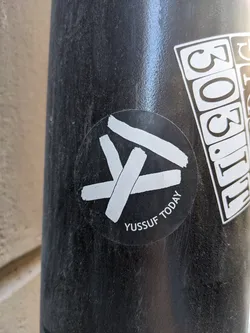 A round sticker with a logo and the text "Yussouf Today". The logo consists of white brushstrokes forming an abstract symbol.