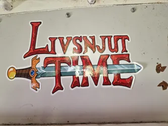 A sticker with the text "Livsnjut Time" written in a stylized font reminiscent of fantasy game titles. A sword is depicted horizontally across the text. The text is predominantly red, with a stone-like texture, and the sword features a gold hilt and a blue jewel.