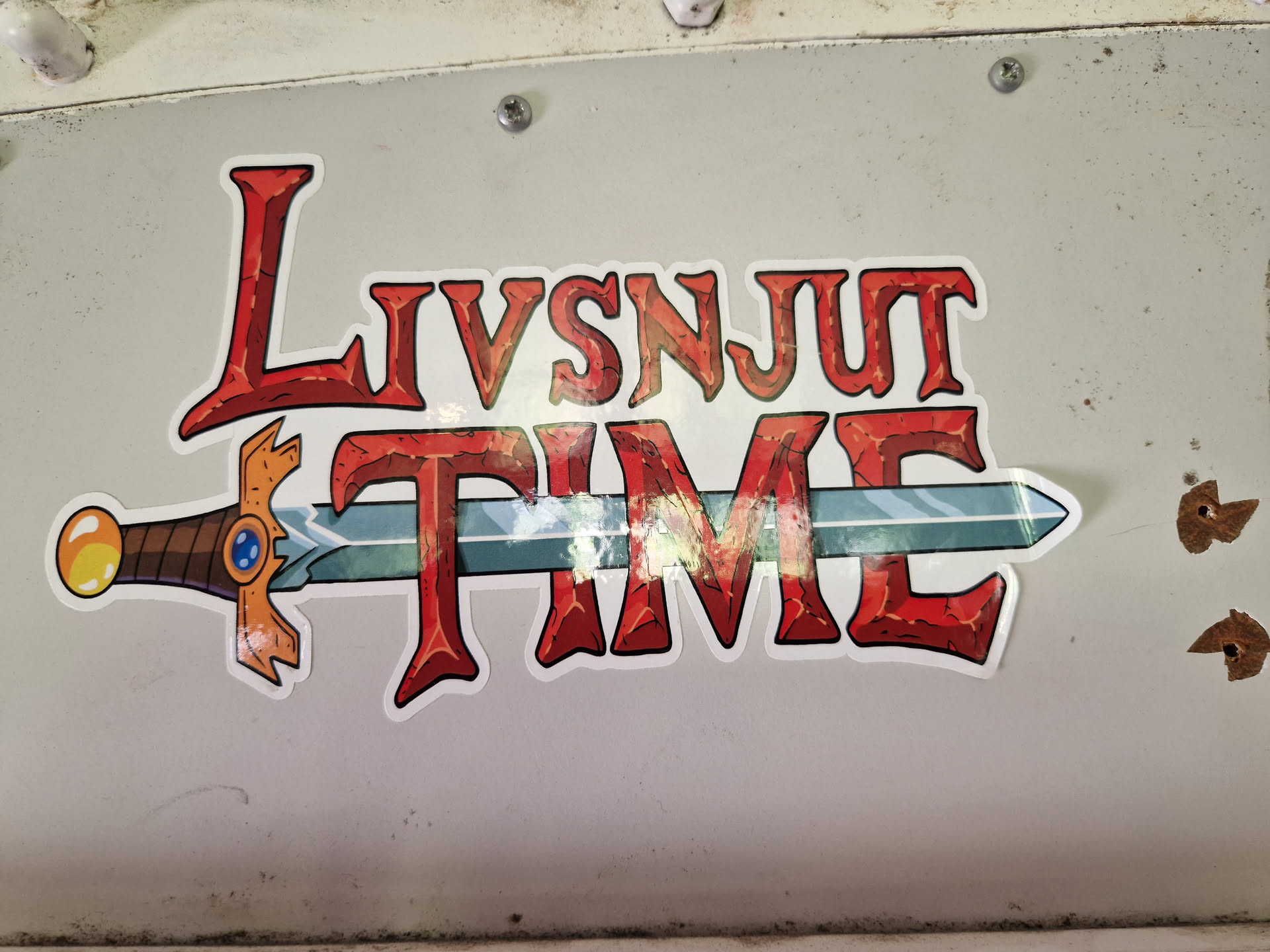 A sticker with the text "Livsnjut Time" written in a stylized font reminiscent of fantasy game titles. A sword is depicted horizontally across the text. The text is predominantly red, with a stone-like texture, and the sword features a gold hilt and a blue jewel.