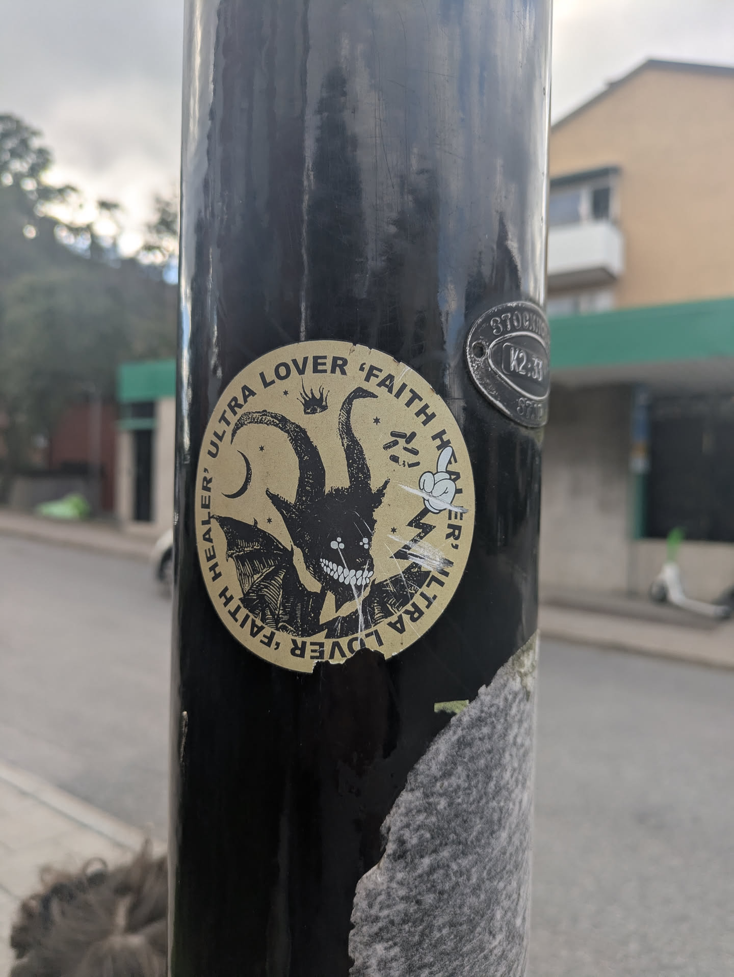 The sticker is circular, featuring a bold black illustration on a muted gold or tan background. The central image is a stylized, horned creature with large, curved horns, bat-like wings, and a wide, toothy grin revealing sharp, white teeth. Its eyes are small, glowing white dots. Above its head, a small crown is visible. To the left, a crescent moon and several small stars are depicted. To the right, a cartoonish white hand with a pointing finger emerges, surrounded by jagged lines resembling lightning bolts. The text "ULTRA LOVER 'FAITH HEALER'" is repeated twice, forming a circular border around the central illustration. The sticker shows some wear, particularly at its bottom edge.