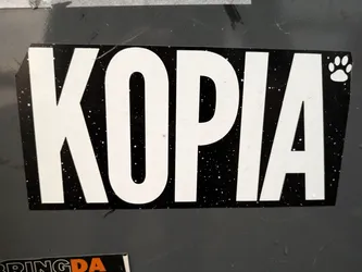 Black and white sticker with the word KOPIA in bold white letters on a black background with white dots, resembling stars. A small paw print is visible in the upper right corner.