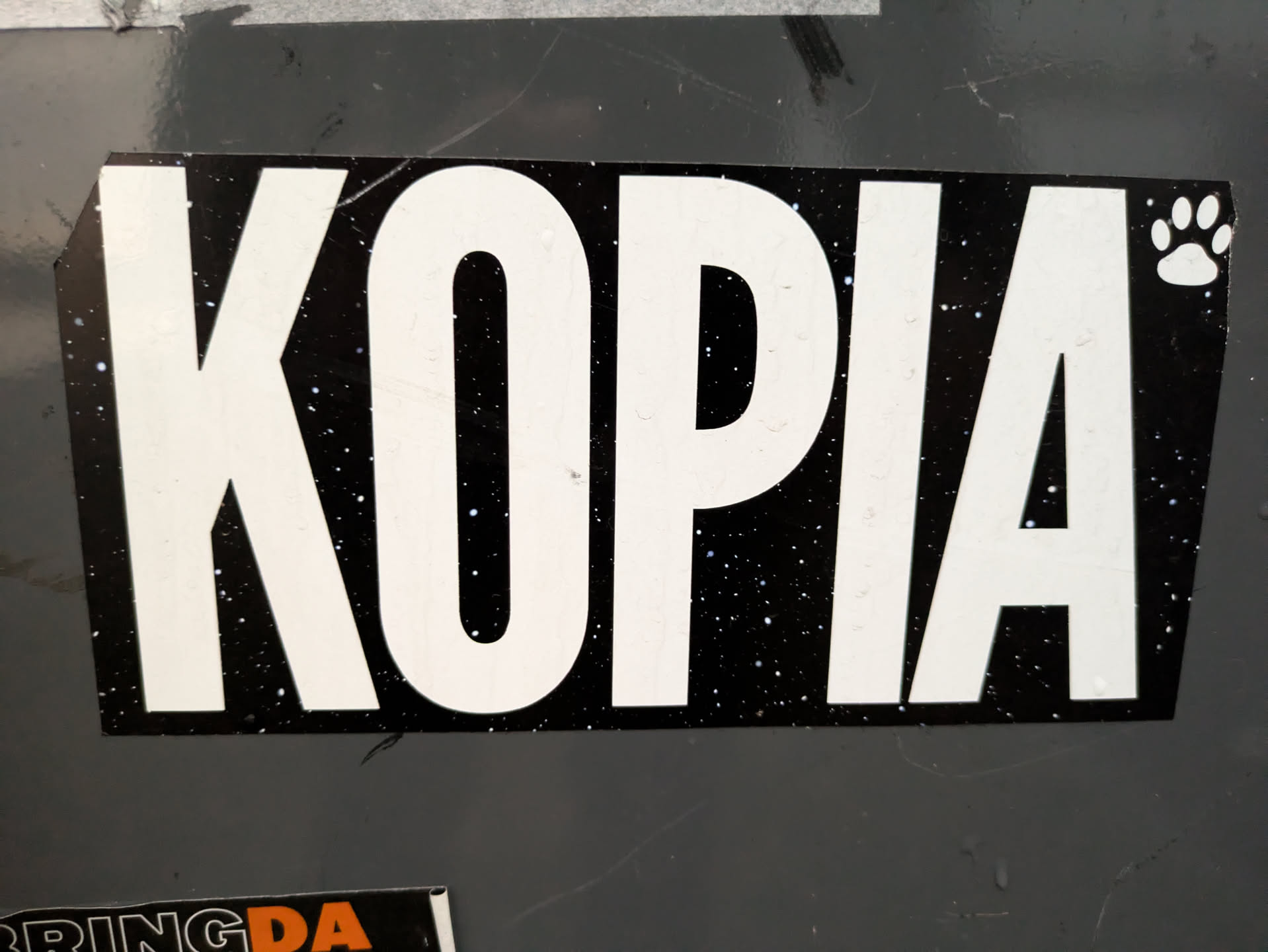 Black and white sticker with the word KOPIA in bold white letters on a black background with white dots, resembling stars. A small paw print is visible in the upper right corner.