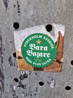 A round green sticker with the text "STOCKHOLM" and "SVERIGE" on top and bottom, with a star in between. In the center, it says "Bara Bajare" in a stylized font. Below it, it says "NON PLUS ULTRA" with two crossed oars between the words. The sticker appears to be pasted on a weathered metal surface.