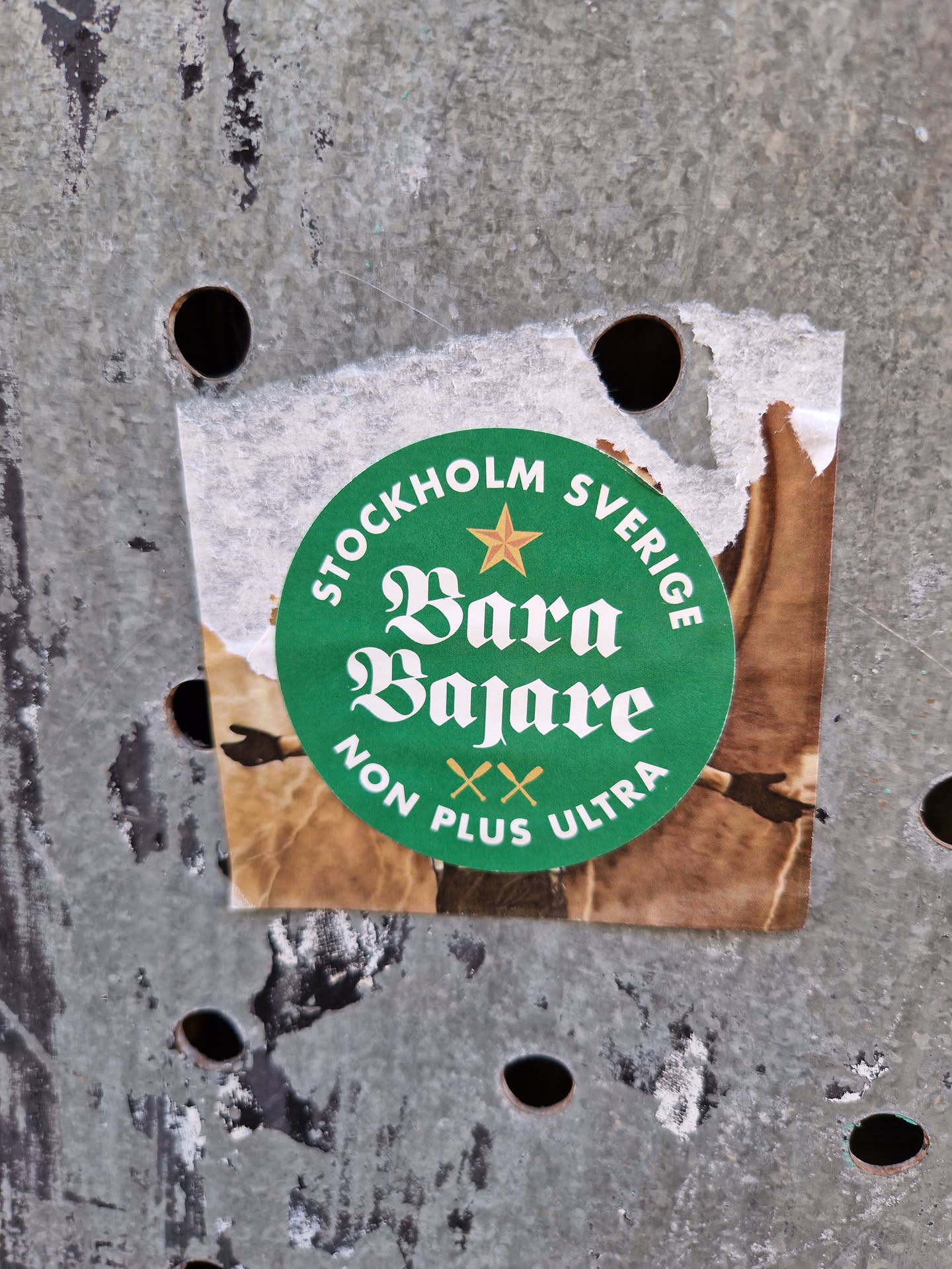A round green sticker with the text "STOCKHOLM" and "SVERIGE" on top and bottom, with a star in between. In the center, it says "Bara Bajare" in a stylized font. Below it, it says "NON PLUS ULTRA" with two crossed oars between the words. The sticker appears to be pasted on a weathered metal surface.