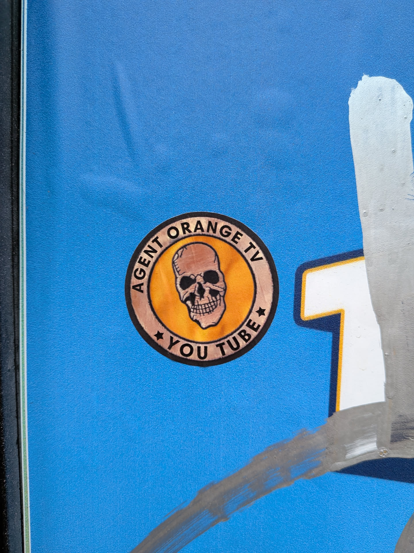 A circular sticker with a vintage aesthetic. It features a central light brown skull with black eye sockets and nose cavity, set against a vibrant orange-yellow background. An outer ring, colored dark brown or black, frames the central image. Text is inscribed on this outer ring: "AGENT ORANGE TV" arches across the top, and "YOU TUBE" is positioned at the bottom, separated by small star symbols on either side. The overall appearance suggests a channel logo or brand identity.