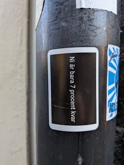 A rectangular sticker with rounded corners and a white border, affixed to a dark metal pole. It features a solid dark background with vertical white text printed reading 'Ni är bara 7 procent kvar' in a modern sans-serif typeface.