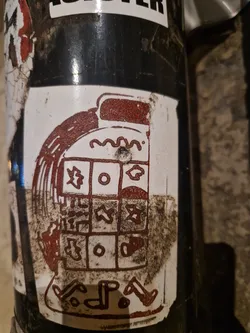 A sticker depicting a stylized machine or device with a grid-like pattern.  The style is reminiscent of ancient symbols or a weathered artifact. The sticker is dirty and shows signs of wear.