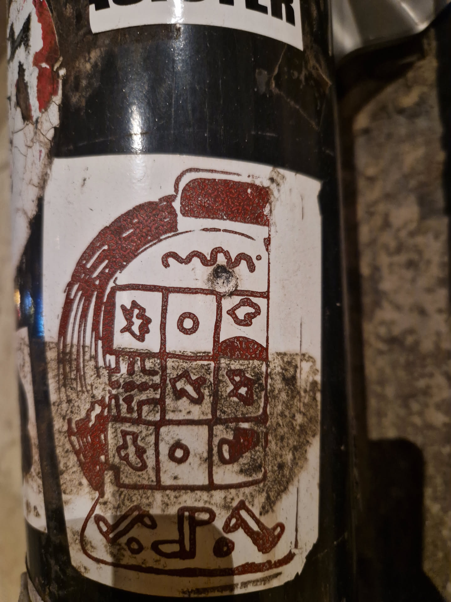 A sticker depicting a stylized machine or device with a grid-like pattern.  The style is reminiscent of ancient symbols or a weathered artifact. The sticker is dirty and shows signs of wear.