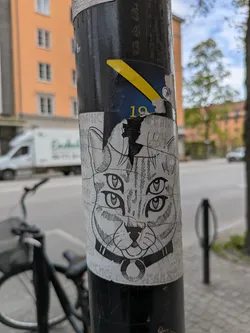 A black and white sticker of a cat's head with four eyes, wearing a collar with a ring. The sticker is applied to a black pole.