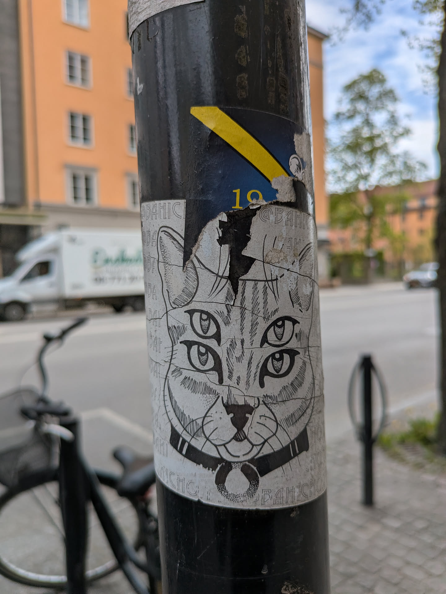 A black and white sticker of a cat's head with four eyes, wearing a collar with a ring. The sticker is applied to a black pole.