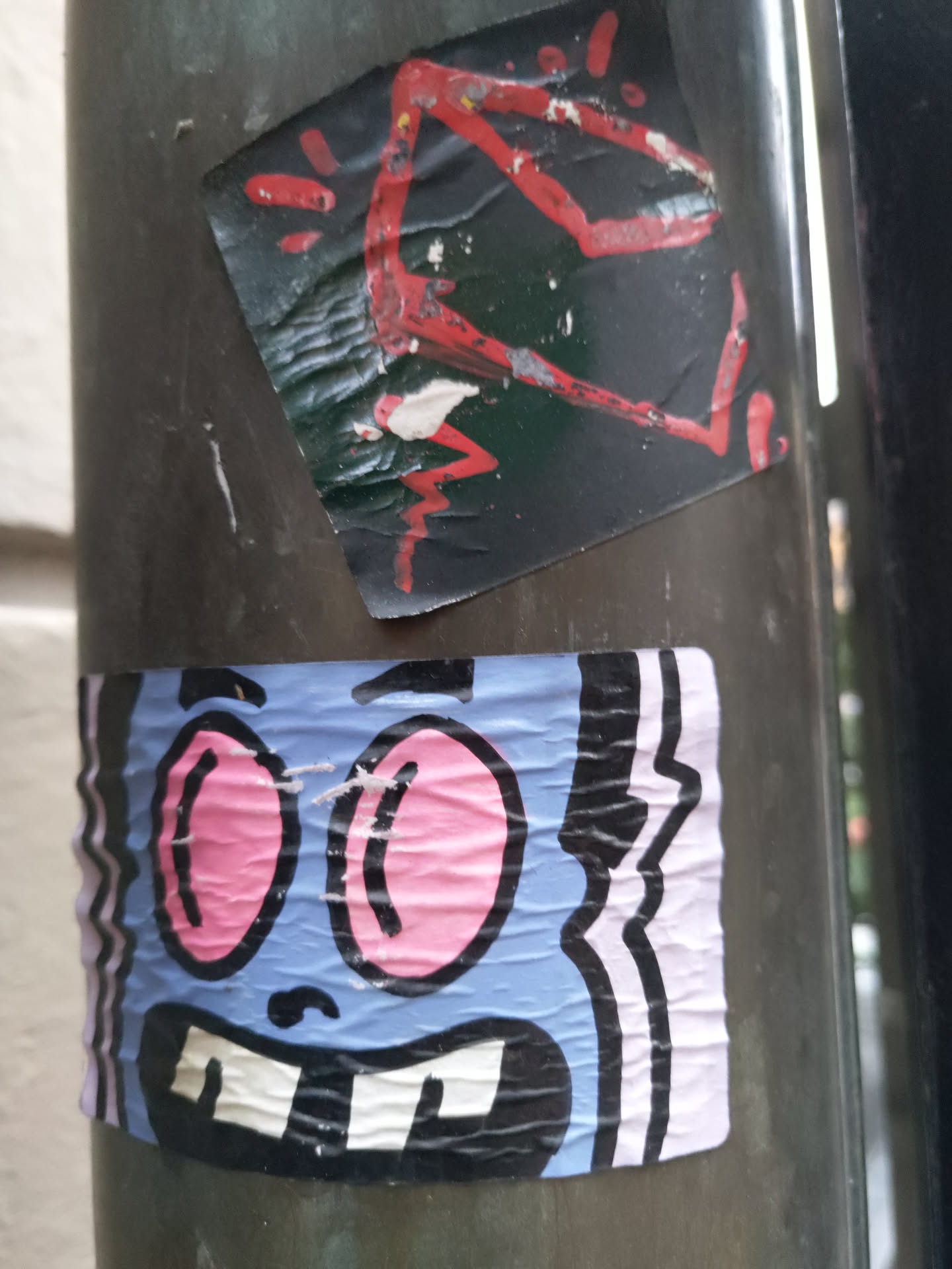 A playful blue cartoon character sticker with large pink eyes and a wide grin. The sticker has a slightly worn look, suggesting it has been exposed to the elements. It is affixed to a dark metal pole. There is another, smaller, more abstract sticker above it.