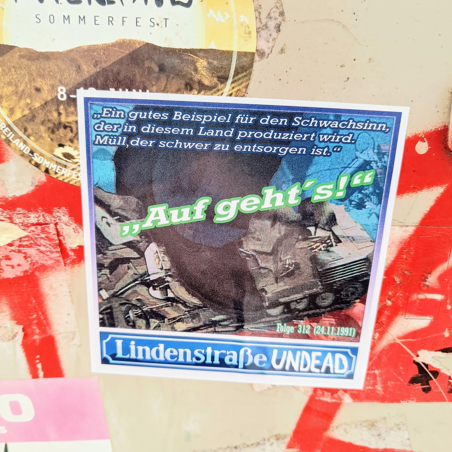 This sticker shows a picture of a tank with the text "Auf geht's!" which means "Let's go!" in German. At the bottom it says "Lindenstraße Undead" and "Folge 312 (24.11.1991)".  A quote about the absurdity of waste production in Germany is included. The overall aesthetic is reminiscent of political or protest art.