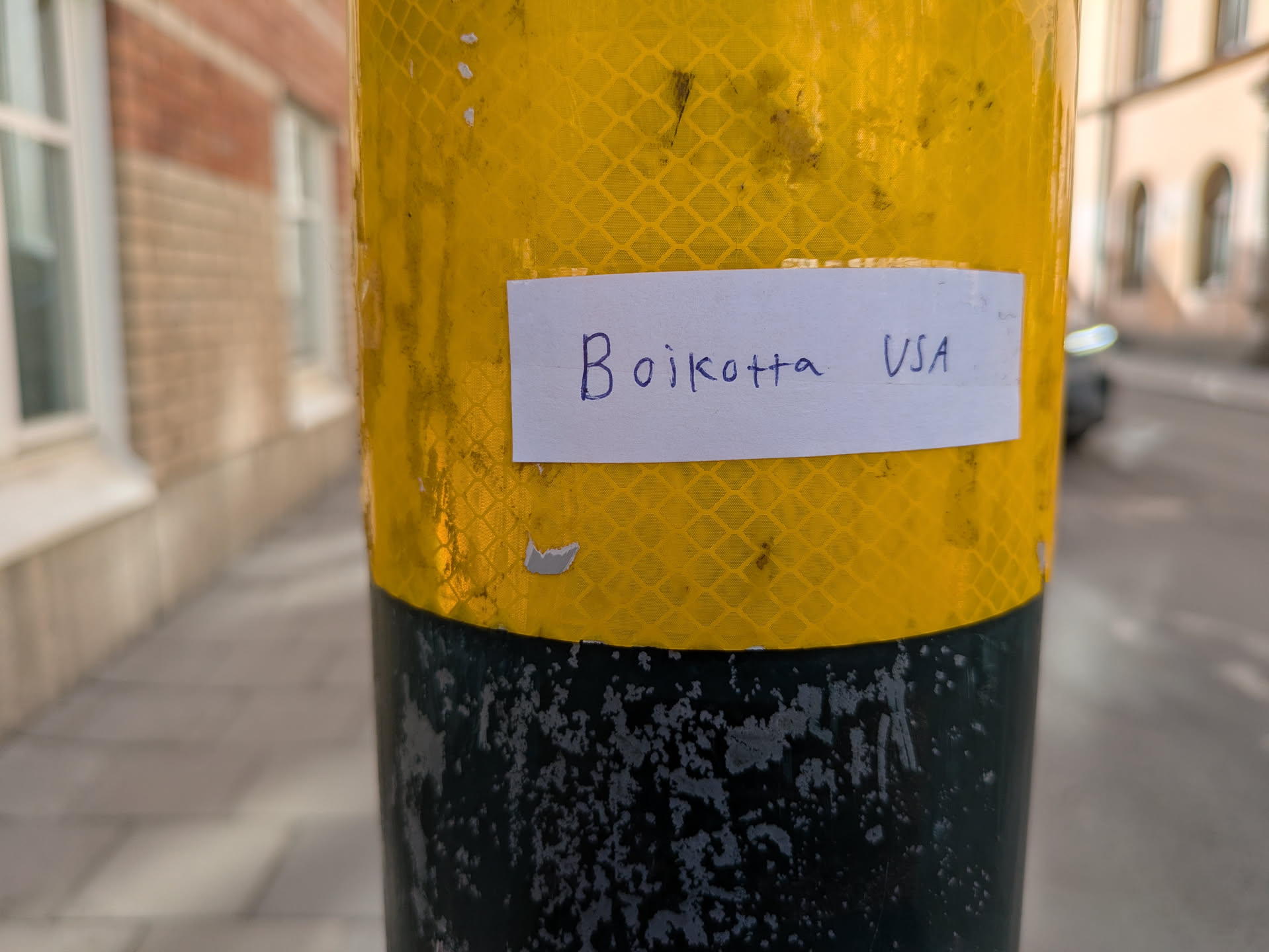 A white rectangular sticker with the text 'Boikotta USA' written in blue ink is stuck to a yellow and black reflective bollard on a street.