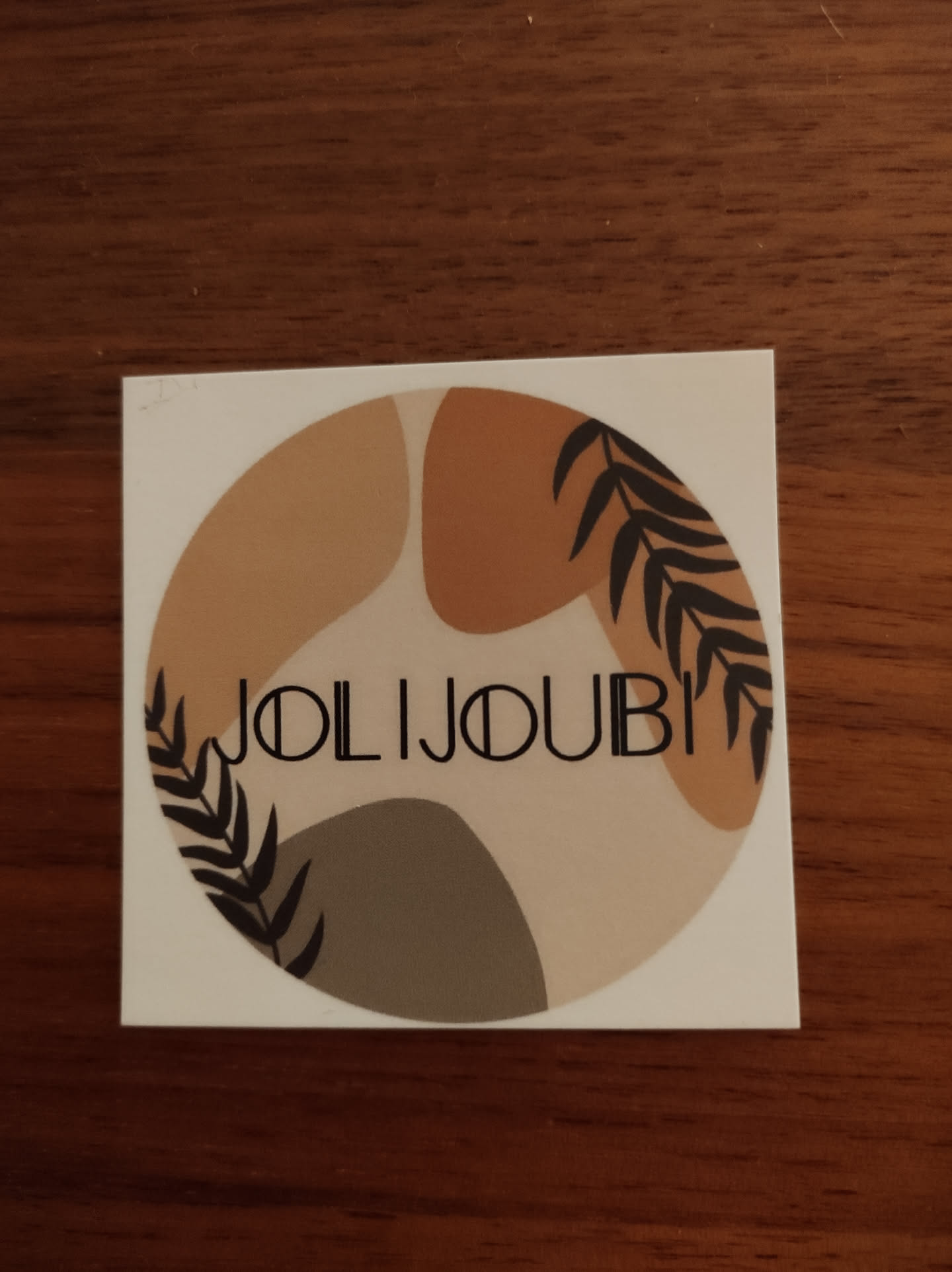 Round sticker with an abstract design in earthy tones.  The sticker features various shades of brown and beige, along with black leaves and the text "JOLIJOUBI".