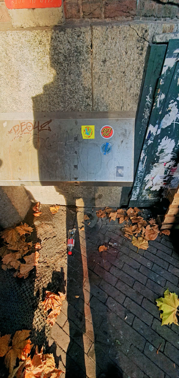 The image features a metal surface adorned with multiple stickers. One sticker reads "JATE DVE". The other stickers are more abstract. The location appears to be an outdoor urban setting, with autumn leaves scattered nearby.