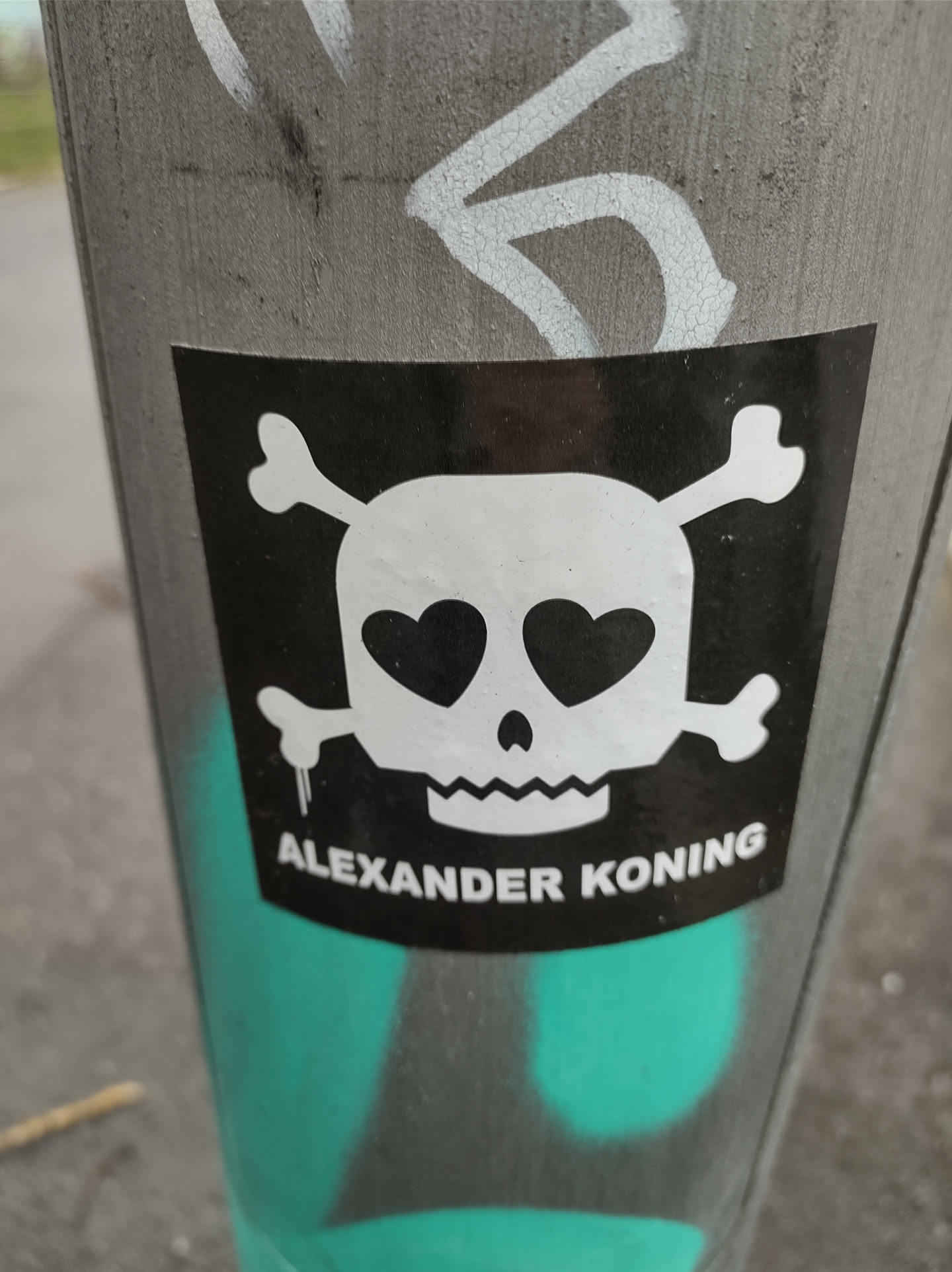 A black and white sticker of a skull with crossbones and heart-shaped eyes. The text 'ALEXANDER KONING' is printed below the skull. The sticker is affixed to a gray pole.