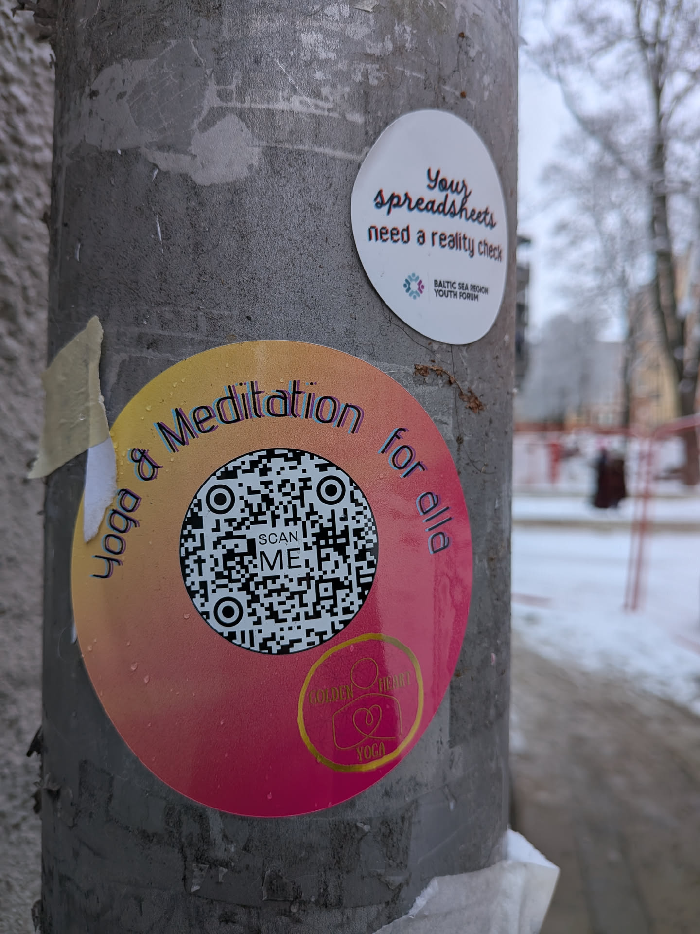 A circular sticker featuring a vibrant gradient background, transitioning from yellow-orange at the top to a deep pink-magenta at the bottom. The sticker is adorned with small water droplets, suggesting outdoor exposure. Around the top arc, the text "Yoga & Meditation for alla" is written in a stylized font with a subtle 3D effect, appearing in shades of blue and purple. A prominent black and white QR code occupies the center, with the words "SCAN ME" embedded within it. At the bottom, a golden outline logo depicts a heart encompassing a figure in a yoga pose, with the text "GOLDEN HEART YOGA" beneath it.