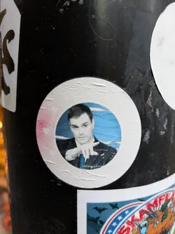 A round sticker featuring a portrait of a man, possibly a character from Rick and Morty, in a formal suit.  The sticker is adhered to a dark surface and shows some slight wear. A small pink heart is visible near the edge.