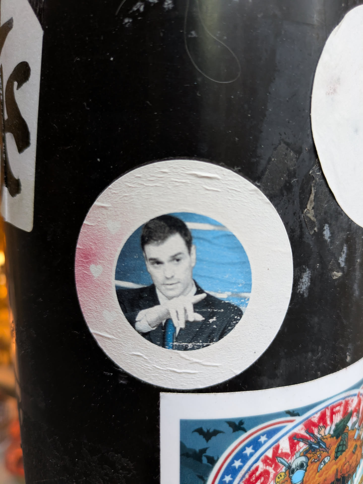 A round sticker featuring a portrait of a man, possibly a character from Rick and Morty, in a formal suit.  The sticker is adhered to a dark surface and shows some slight wear. A small pink heart is visible near the edge.
