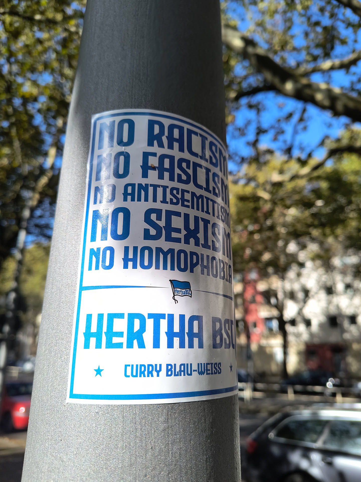A sticker featuring the Hertha BSC logo and the message 'No Racism, No Fascism, No Antisemitism, No Sexism, No Homophobia'. The sticker is affixed to a lamppost.