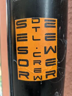 A close-up shot of an orange sticker with the word "SCREWDRIVER" written in a stylized font. The sticker is adhered to a black cylindrical surface. The letters are arranged in a visually interesting way, almost like a puzzle.