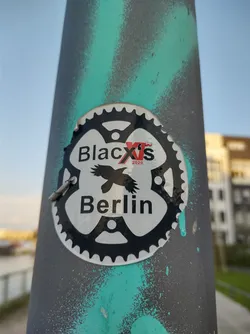 A sticker featuring a black and white bicycle chainring design with a raven and the text 'BlackXs 2020' and 'Berlin'. It's affixed to a teal and gray pole.