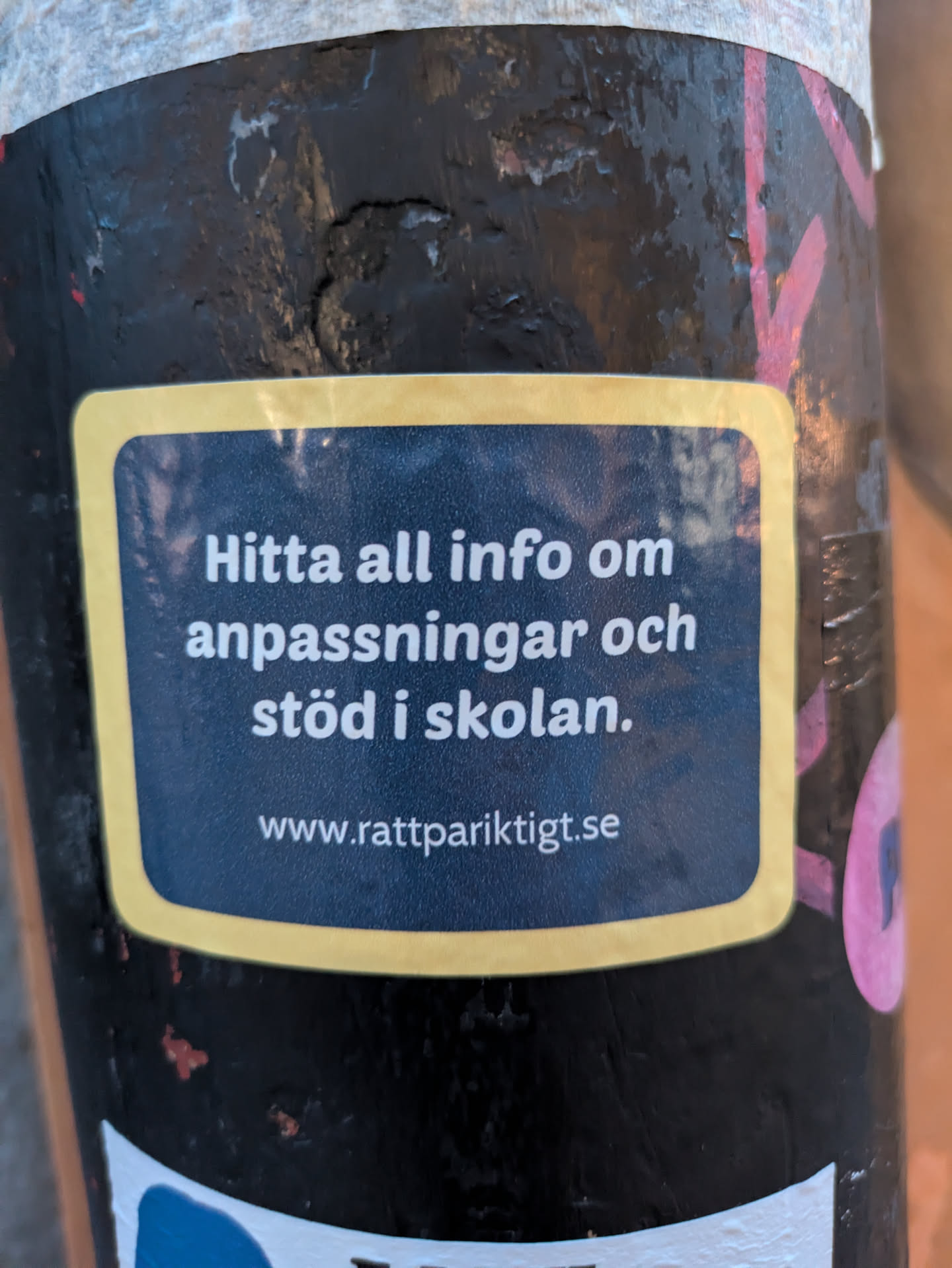 The sticker is rectangular with rounded corners and a yellow border. The background of the sticker is dark blue, almost black, with a subtle textured pattern.  White text is printed on the dark blue background. The top line reads 'Hitta all info om'. The second line reads 'anpassningar och'. The third line reads 'stöd i skolan.'. Below this, centered, is a website address: 'www.rattpariktigt.se'. The sticker appears to be adhered to a dark, textured surface, possibly a pole, with some wear and tear visible on the surrounding material.