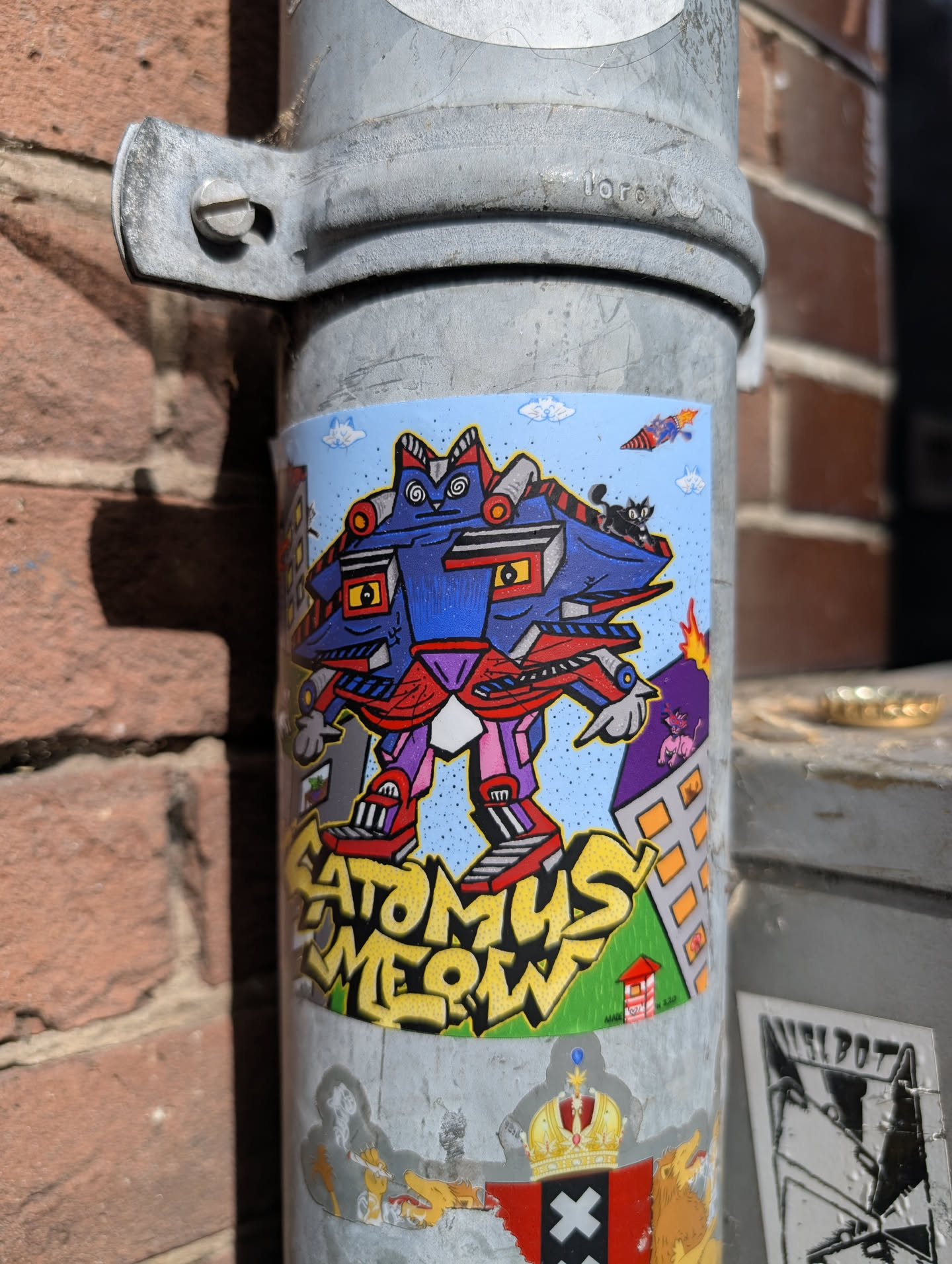 The sticker showcases a whimsical robot with cat-like features, standing tall over a stylized cityscape with cartoonish details like a volcano. The text "ATOMUS MEOW" is present. The style is cartoonish and colorful.