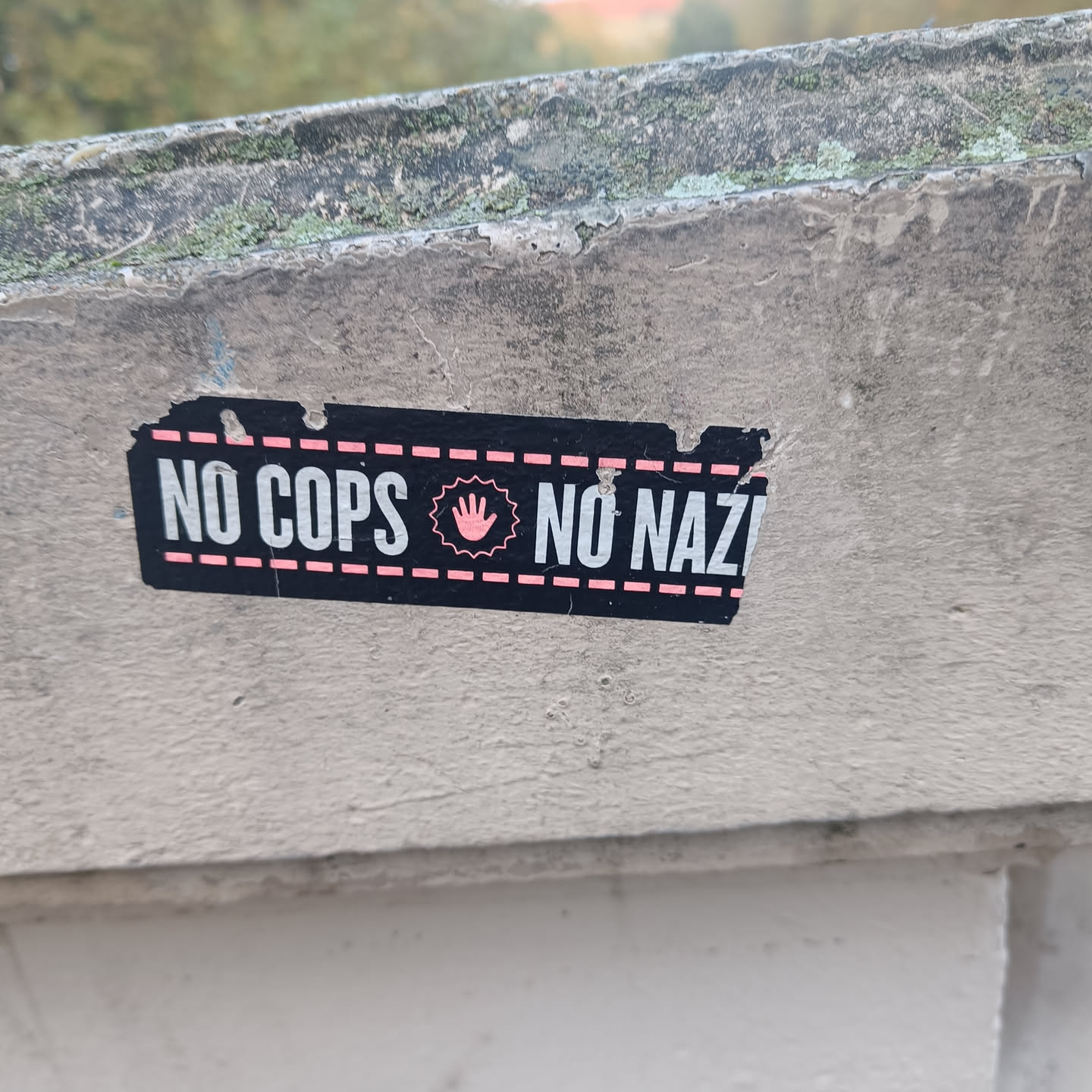 A black and pink sticker with the text "NO COPS NO NAZIS" and a pink hand symbol in the center. The sticker is attached to a light grey wall.