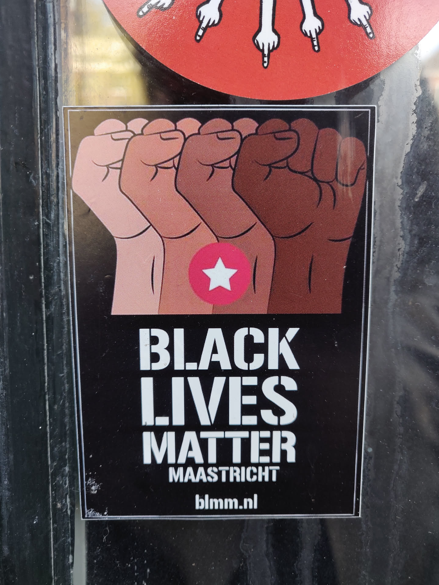 A Black Lives Matter sticker featuring four fists of different skin tones raised in solidarity.  The sticker has a pink star and the website blmm.nl.