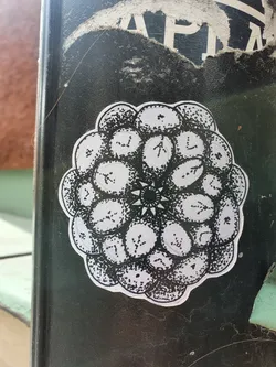 A round sticker with a detailed drawing in black ink on a white background. The design is composed of multiple circular elements arranged in a concentric pattern, resembling a blackberry or a flower. Each circular element contains small, intricate details, and the center features a smaller, star-like motif. The style is reminiscent of a mandala or a botanical illustration.