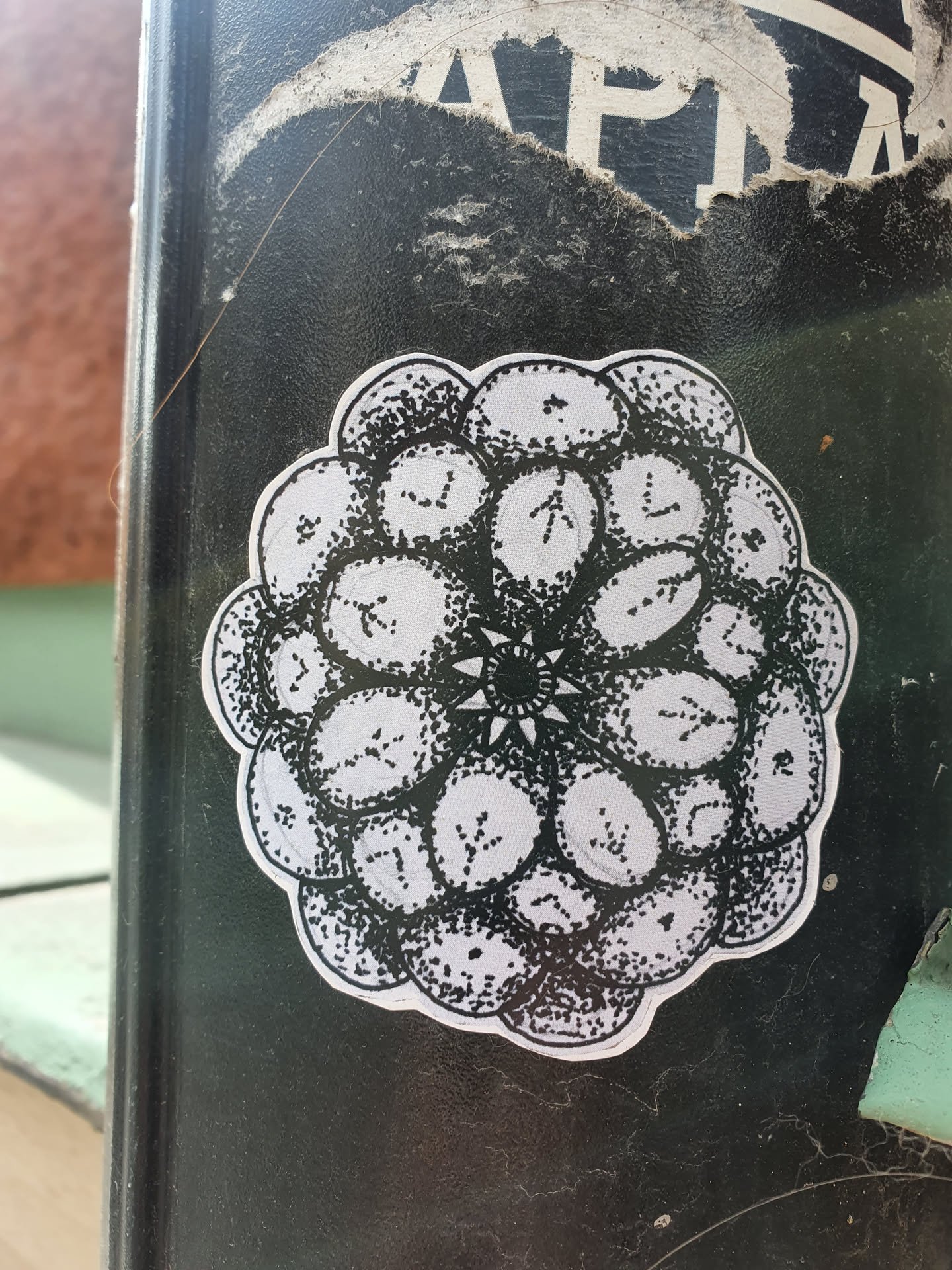 A round sticker with a detailed drawing in black ink on a white background. The design is composed of multiple circular elements arranged in a concentric pattern, resembling a blackberry or a flower. Each circular element contains small, intricate details, and the center features a smaller, star-like motif. The style is reminiscent of a mandala or a botanical illustration.