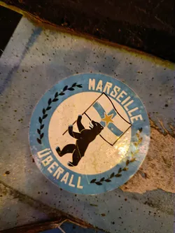 A round sticker with a blue background and a white center depicts a black bear standing on its hind legs, raising a light blue and white flag with a yellow star.  The text "MARSEILLE" is written in a circular arc at the top, and "ÜBERALL" is written in a similar arc at the bottom. The text is enclosed in a wreath of olive branches.