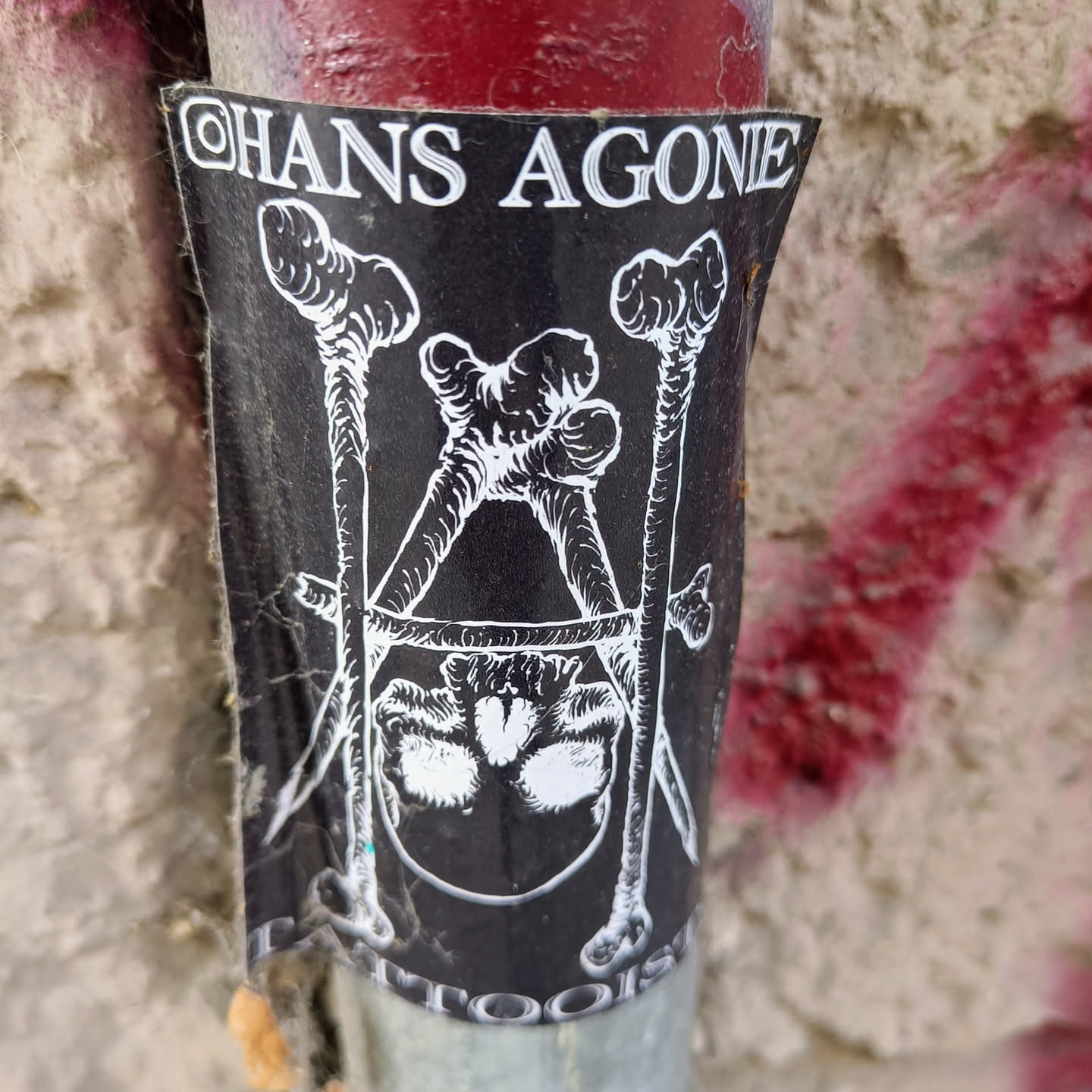 A black and white sticker with an illustration of a skull bound by bones. The text "Hans Agone" is visible at the top. It appears to be a tattoo artist's sticker.