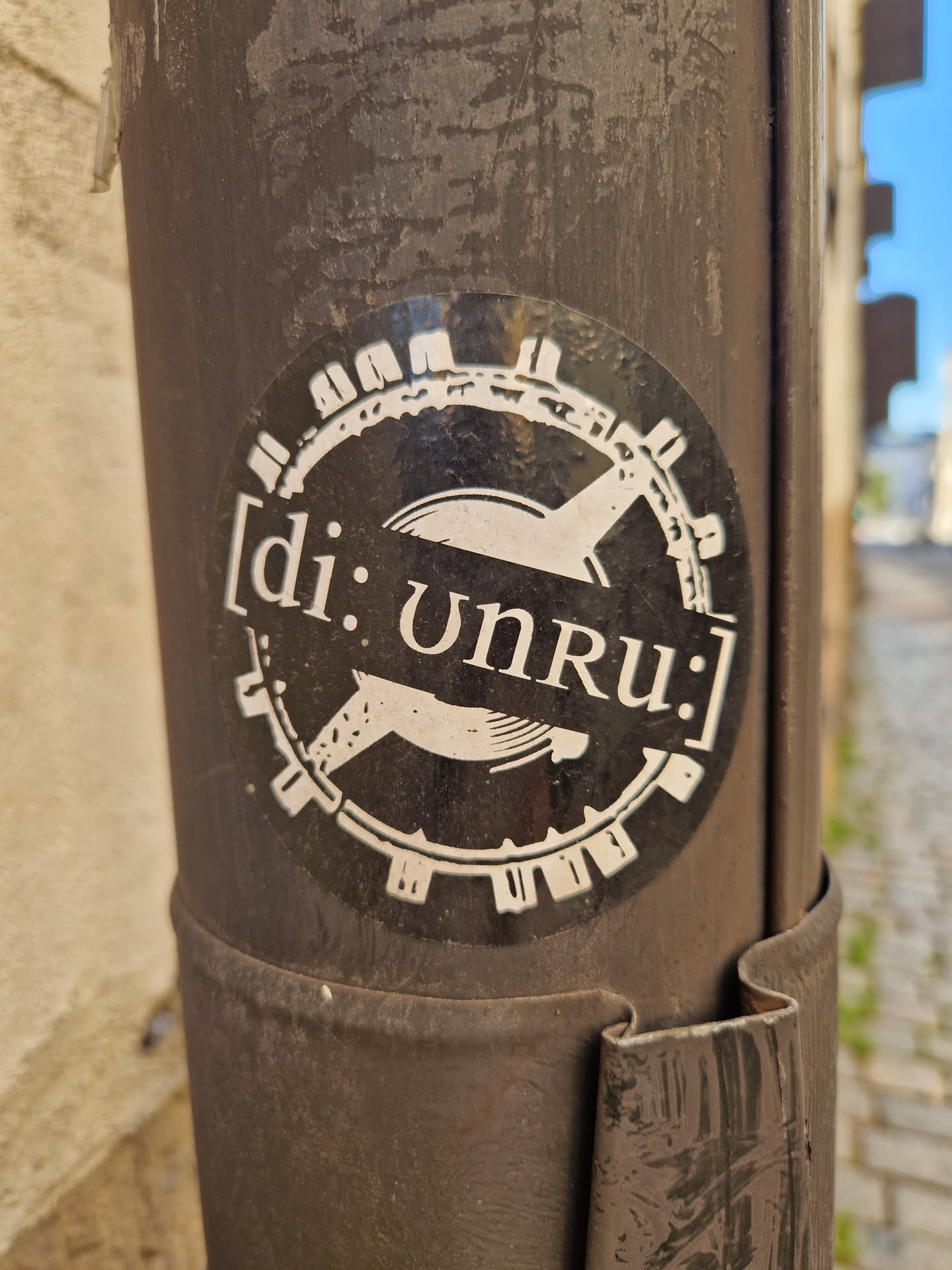 A black and white circular sticker with the text [di:Unru:] in the center. The sticker is attached to a dark brown pole.
