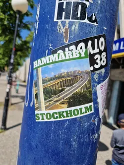 A sticker featuring a picture of a highway overpass in Hammarby, Stockholm. The sticker is attached to a blue pole.