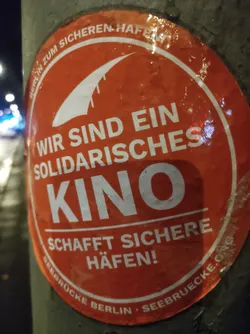 A round orange sticker with white text and a white graphic of a boat. The text translates to "We are a solidarity cinema. Create safe harbors!"  The bottom also includes the website address seebruecke.org and the city Berlin.