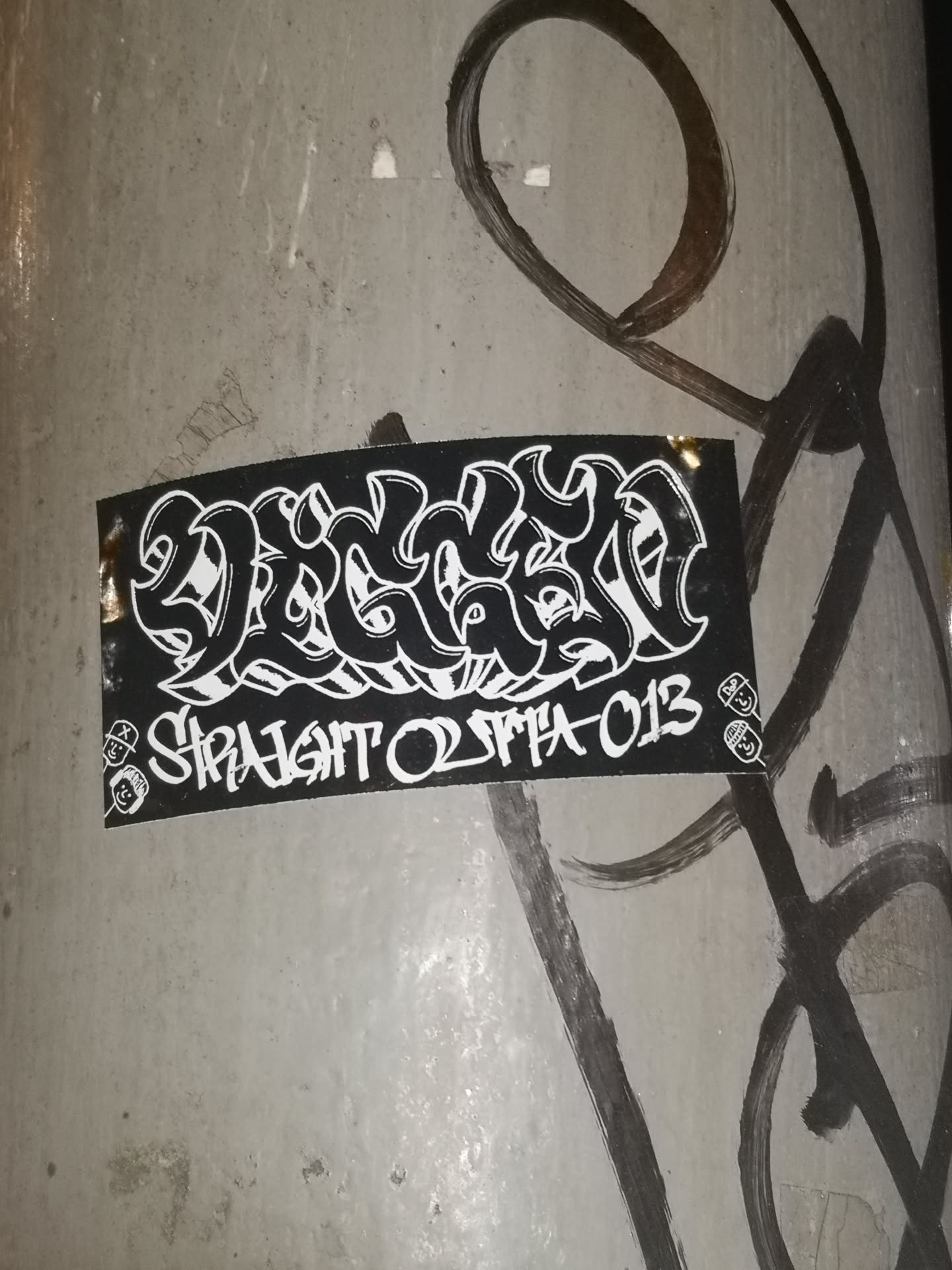 A rectangular black sticker featuring prominent white graffiti lettering. The main text, 'DRAGON', is rendered in a highly stylized, intricate wildstyle or bubble-letter font, filling the upper portion of the sticker. Below this, in a smaller, blockier but still stylized white font, is the phrase 'STRAIGHT OUTTA 013'. On both the left and right sides, flanking the lower text, are small, simple white cartoon faces stacked vertically; the top face wears a cap, and the bottom face wears headphones. The sticker has a slight sheen and is adhered to a textured, light grey surface, possibly a concrete pillar, with some dark, abstract graffiti strokes visible around it.
