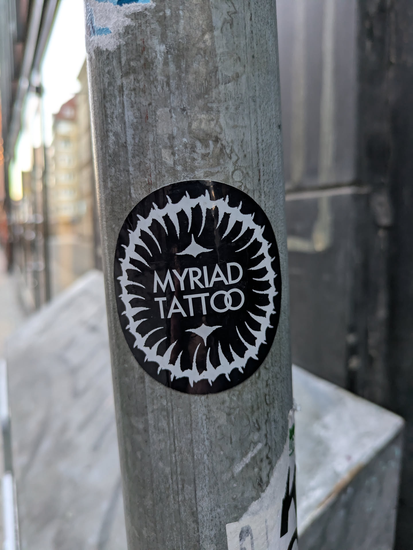 This is a black and white circular sticker adhered to a gray metal pole. The sticker features the words "MYRIAD TATTOO" in the center, surrounded by a sunburst-like design composed of white teardrop shapes pointing inwards.